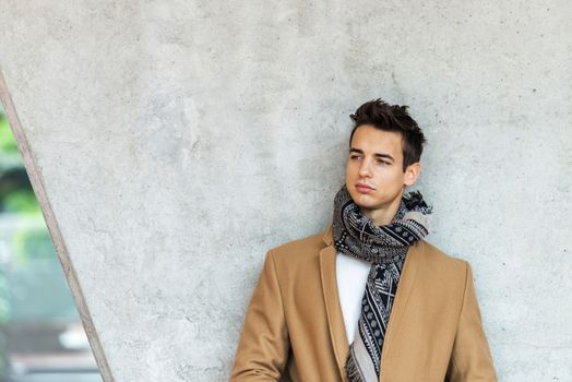 Front view of stylish young man wearing coat and scarf leaning on a wall while looking away