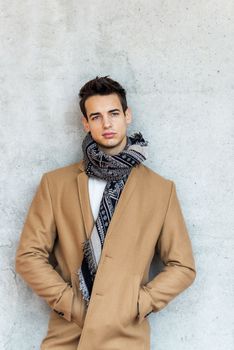 Front view of stylish young man wearing coat and scarf leaning on a wall while looking camera