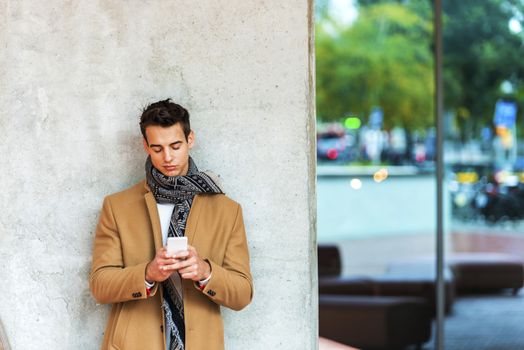 Front view of fashionable young man wearing denim clothes leaning on a wall while using a mobile phone outdoors