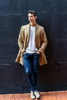 Front view of a trendy young man standing against black wall while looking away outdoors in the street