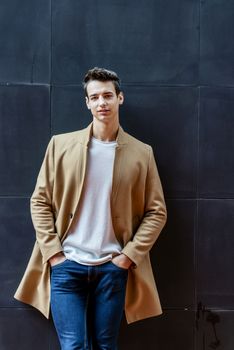 Front view of a trendy young man standing against black wall while looking camera outdoors in the street