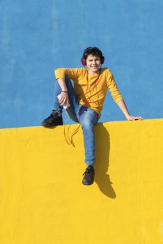 Front view of a young boy wearing casual clothes sitting on a yellow fence against a blue wall while using a mobile phone to listening music