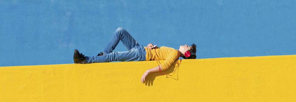 Front view of a young boy wearing casual clothes lying on a yellow fence against a blue wall while using a mobile phone to listening music