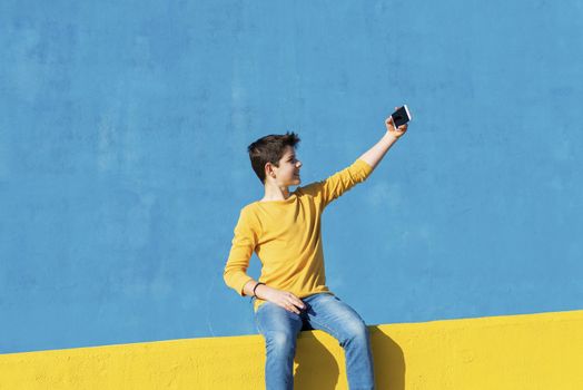 Front view of a young boy wearing casual clothes sitting on a yellow fence against a blue wall while using a mobile phone