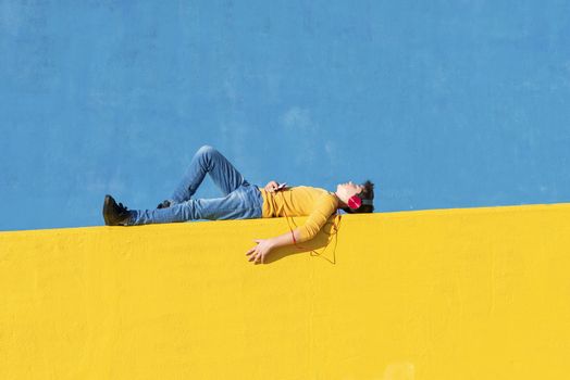 Front view of a young boy wearing casual clothes lying on a yellow fence against a blue wall while using a mobile phone to listening music