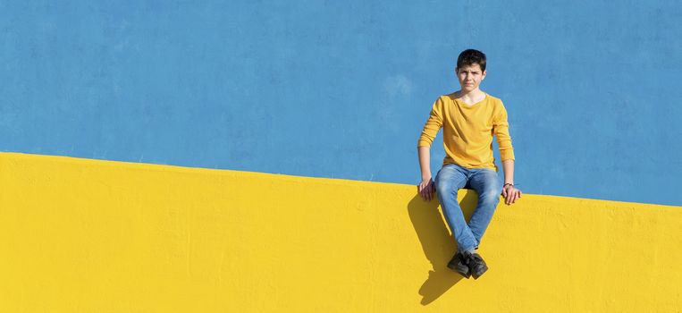 Front view of a young boy wearing casual clothes sitting on a yellow fence against a blue wall