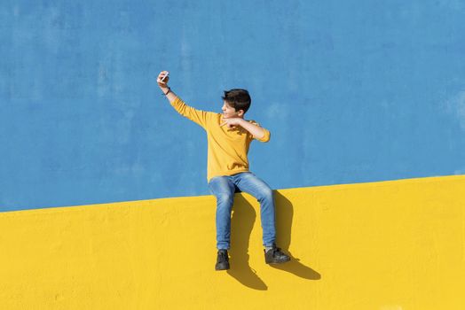Front view of a young boy wearing casual clothes sitting on a yellow fence against a blue wall while using a mobile phone