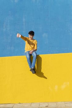 Front view of a young boy wearing casual clothes sitting on a yellow fence against a blue wall while using a mobile phone