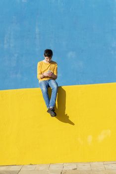 Front view of a young boy wearing casual clothes sitting on a yellow fence against a blue wall while using a mobile phone