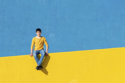 Front view of a young boy wearing casual clothes sitting on a yellow fence against a blue wall
