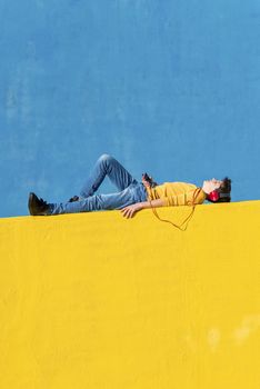 Front view of a young boy wearing casual clothes lying on a yellow fence against a blue wall while using a mobile phone to listening music