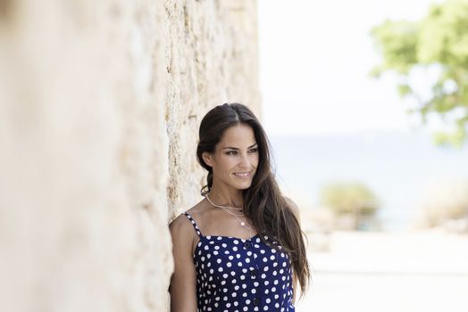 Beautiful smiling woman in blue dress leaning on wall while looking away