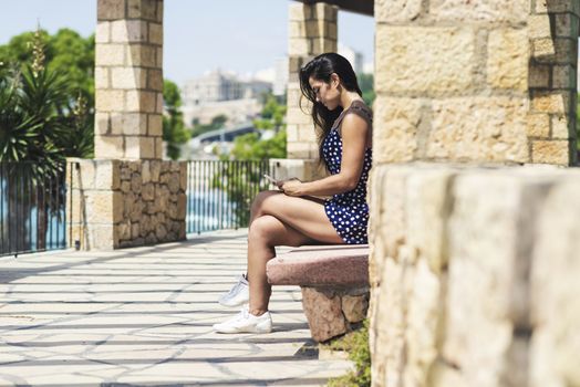 Beautiful woman in blue dress sitting on a bench while using a smartphone