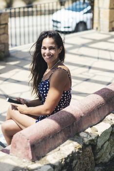 Top view of a smiling woman in blue dress sitting on a bench while using a mobile phone while looking to camera outdoors