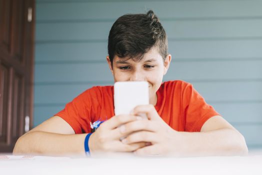 Handsome Caucasian 13-year old boy wearing red t-shirt sitting outdoors using electronic gadget. Schoolboy holding mobile phone, messaging friends