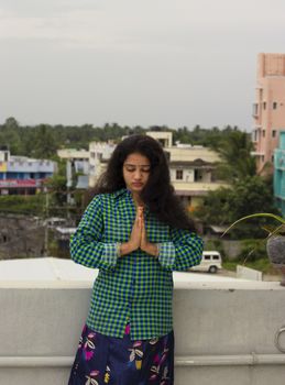 A young Hindu model standing in an open environment above the roof of the house and closing the eyes and praying with his hands to the Sun God above the head and wearing a green shirt.
