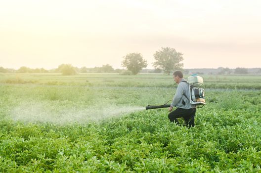 Farmer with a mist sprayer blower processes the potato plantation. Protection and care. Environmental damage and chemical pollution. Use of industrial chemicals to protect crops from insects and fungi