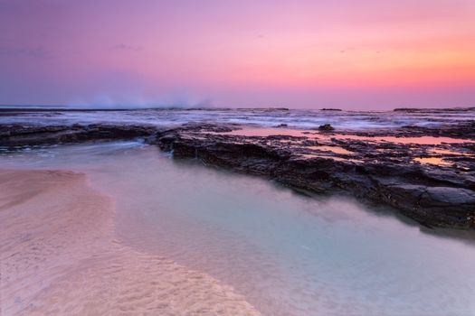 Pretty sunrise of pink, mauve and orange over the beach and rock shelf with reflections