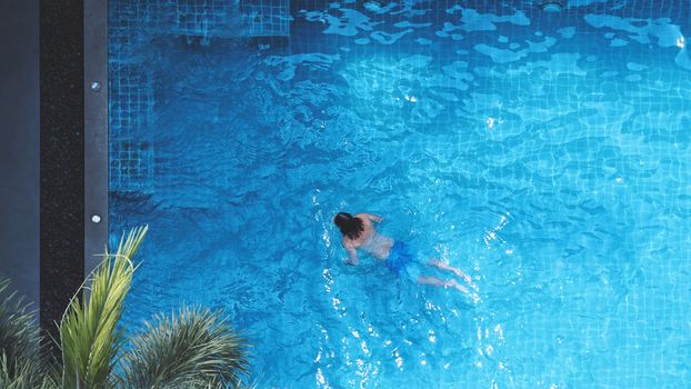 Swimming pool top view angle which young man relaxing and enjoying in pools of his hotel in summer season of Thailand at vacation day and have sun light and flares on blue water surface.