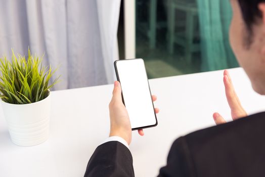 Work from home, Back view of Asian young businessman smile wearing suit video conference call or facetime by smart digital mobile phone blank screen raise hand to say hello teammates on desk