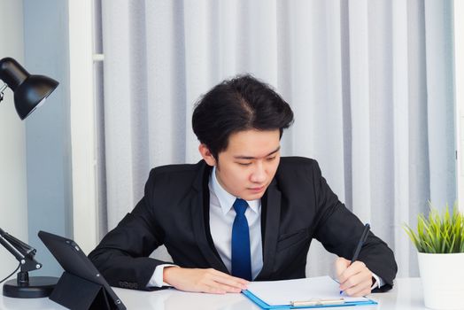 Work from home, Asian young businessman video conference call or facetime he smiling looking to paper sitting on desk using smart digital tablet computer and write notebook information at home office