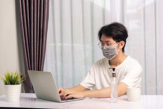 Asian business young handsome man glasses, wearing protective mask with disinfectant gel beside he work from home office remote online with laptop computer quarantines disease coronavirus or COVID-19