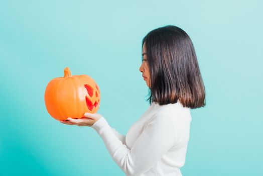 Portrait of Asian beautiful young woman holding orange model pumpkins and looking it, funny happy female with ghost pumpkins, studio shot isolated on blue background