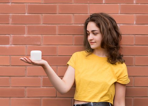 young woman wearing yellow shirt, looking at tub of cream against brick wall