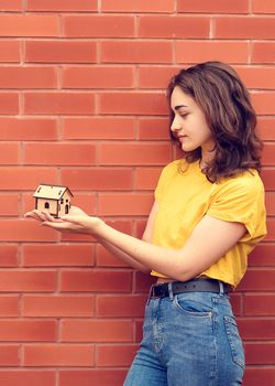 young woman wearing yellow shirt, looking at tub of toy wooden house against brick wall