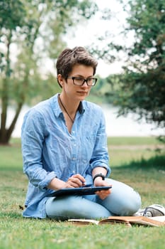 The woman sitting on green grass and studying online on tablet