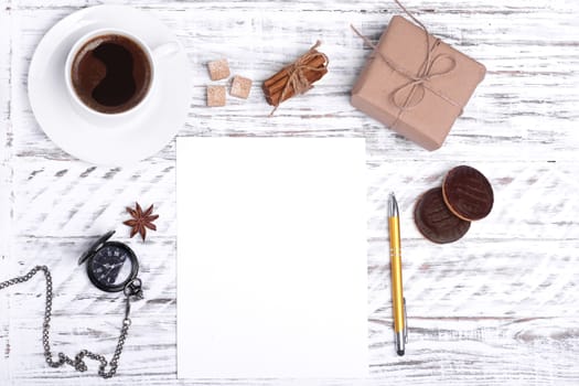 Mockup of empty paper, a white cup of coffee, pen, cake, sugar, anise and gift on white wood desk. Flat Lay with no people. View from the top.