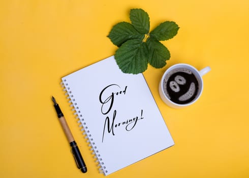 A cup of coffee with smile, notebook with the sign, ink pen and leaf on yellow background
