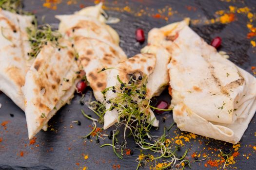 Lavash with cheese on a flat black plate decorated with micro green and pomegranate seeds. Restaurant dish.