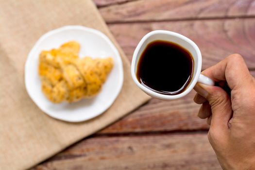 Breakfast concept. Hand holding white cup of coffee with blurry croissants background. and wooden table. 