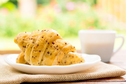 Breakfast concept. Croissants with perilla seeds on white plate and white cup of black coffee on wooden table with green bokeh background.