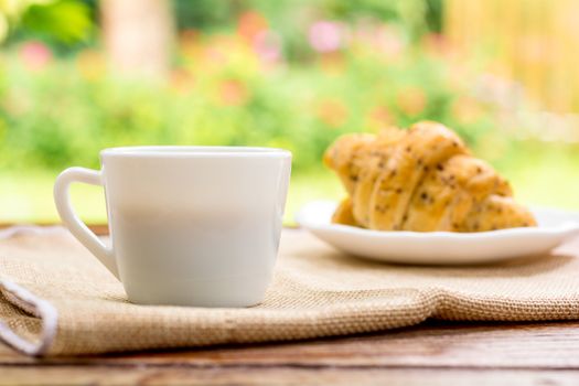 Breakfast concept. White cup of coffee with blurry croissants background on wooden table. 