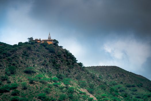 hindu temple on a hill covered in vegetation with the tower and spire clearly visible in the distance with a background of dark monsoon clouds shot in Rajasthan India. Shows the beautiful landmark locations used in the past to build temples and the amazing views from them