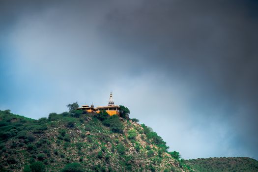 hindu temple on a hill covered in vegetation with the tower and spire clearly visible in the distance with a background of dark monsoon clouds shot in Rajasthan India. Shows the beautiful landmark locations used in the past to build temples and the amazing views from them