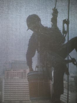 Silhouette images and shoot through transperent curtain effect from inside building which a man cleaning the window of high office building with his equipment such as wipe, sponge, bucket and high risk of danger