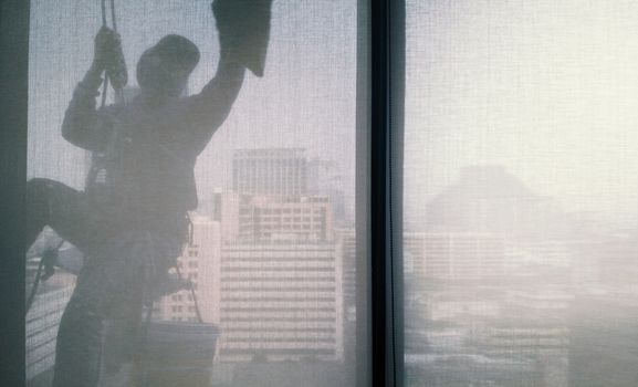 Silhouette images and shoot through transperent curtain effect from inside building which a man cleaning the window of high office building with his equipment such as wipe, sponge, bucket and high risk of danger