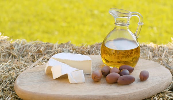 Close up olive oil in a glass jug, cheese and olives on the wooden cutting board in the garden at summer day. Healthy lifestyle concept