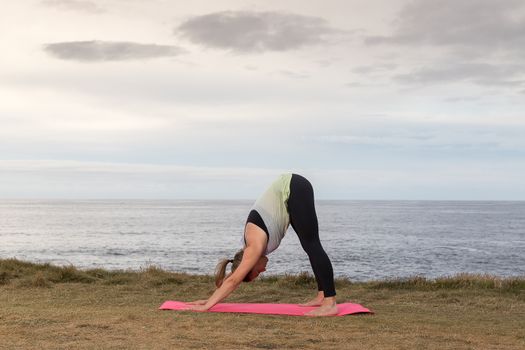 Woman in sportswear doing yoga outdoor on a pink mat with the sea in the background.