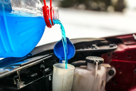 Detail on pouring antifreeze liquid screen wash into dirty car from blue and red anti freeze water container.