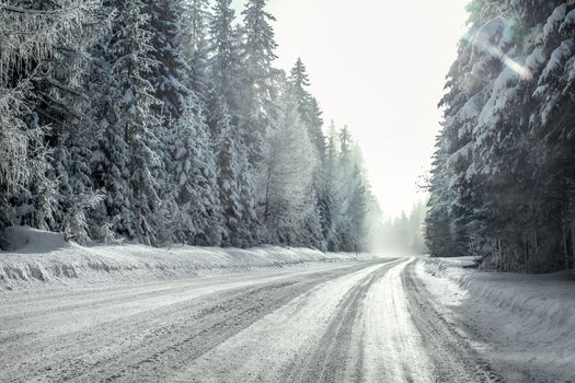 View from a car riding through snow covered winter road curve, lit by strong sun backlight and fog and haze in distance - dangerous driving conditions.