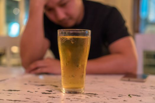 Asian young man in lonely and depressed action and holding head in hands and see glass of beer in pub and restaurant with low light place, depression and drinking concept