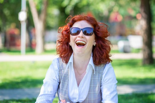 Cheerful girl sits on the grass in the park and laughs out loud. Beautiful girl with red curly hair in sunglasses