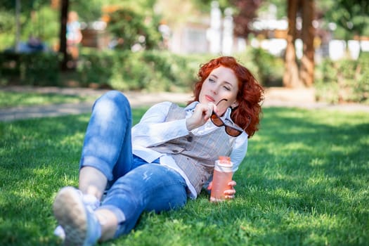 A cute red-haired girl lies in the park on the grass and thinks or dreams about something. Woman walking in a summer park with a bottle of water