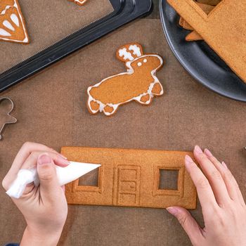 Young woman is decorating Christmas Gingerbread House cookies biscuit at home with frosting topping in icing bag, close up, lifestyle.