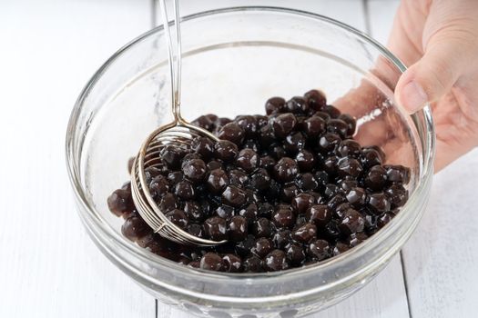 Making bubble tea, scoop and pour cooked brown sugar flavor tapioca pearl bubble balls into cup on white wooden table background, close up, copy space.