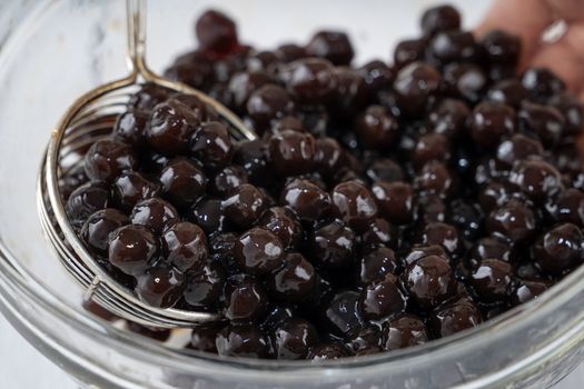 Making bubble tea, scoop and pour cooked brown sugar flavor tapioca pearl bubble balls into cup on white wooden table background, close up, copy space.